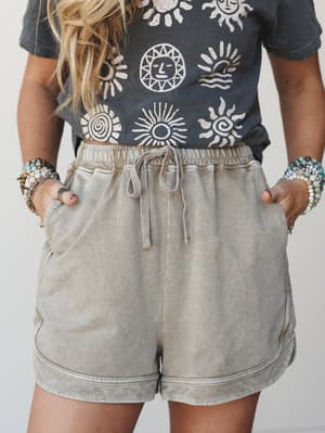 A mid-shot of acid-washed lounge shorts in warm mocha paired with a charcoal sun-print graphic tee, hands tucked in pockets and stacked beaded bracelets adding that effortlessly collected boho finish.