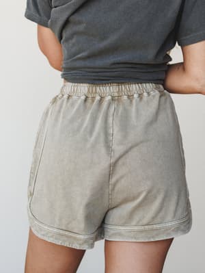 A close mid-shot from behind showing acid-washed lounge shorts in a warm stone-mocha tone, paired with a washed charcoal tee — relaxed, lived-in, and the kind of outfit that makes staying home feel intentional.