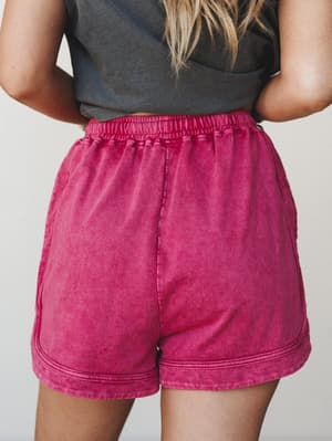 A close-up back view of acid-washed fuchsia lounge shorts with a smocked elastic waistband, paired with a charcoal graphic tee — relaxed, lived-in, and the kind of comfy you don't want to change out of.