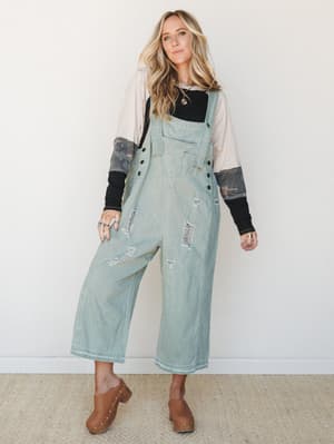 Wide-leg distressed denim overalls in a faded sage wash, layered over a mixed-print long sleeve top and finished with tan studded clogs — effortlessly lived-in and the kind of outfit that looks like you've had it forever.