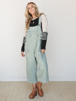 Light sage-washed denim overalls styled over a layered long-sleeve top, worn with brown clogs and stacked jewelry — effortlessly lived-in and the kind of outfit you'd actually want to wear on a Saturday.