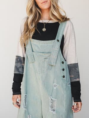 A layered, effortlessly lived-in look built around light sage distressed denim overalls worn over a color-blocked long sleeve with camo sleeves — the kind of outfit that looks like you tried just the right amount.