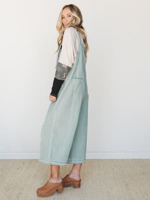 A light-wash sage denim overall jumper styled over a layered long-sleeve tee and camo underlayer, paired with brown wooden clogs — the kind of effortless, thrown-together look that somehow always works.