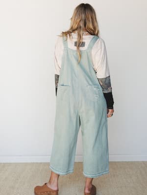 Shot from the back, she's wearing light wash wide-leg denim overalls layered over a blush long-sleeve tee and a printed undershirt, styled with tan studded clogs — the kind of effortless, lived-in look that feels just as at home at the farmers market