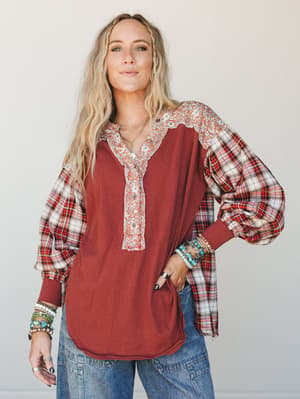 A relaxed mixed-print top in deep rust combines a solid knit body with plaid balloon sleeves and a floral placket, styled with wide-leg jeans and stacked turquoise bracelets — the kind of top that looks like it was meant to be worn with coffee in han