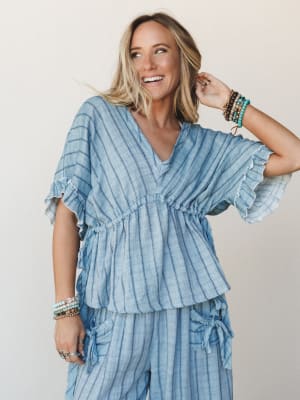 A laughing blonde model wears a matching striped linen set — a breezy v-neck blouse with flutter sleeves and a drawstring waist paired with wide-leg pants — layered with stacks of beaded bracelets against a warm neutral studio backdrop.