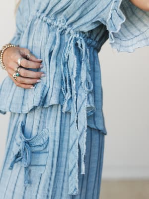 A close-up detail shot of a chambray striped set — the frayed tie waist, ruffle hem, and layered texture do all the talking, styled with stacked silver rings and a turquoise stone for that effortless boho finish.