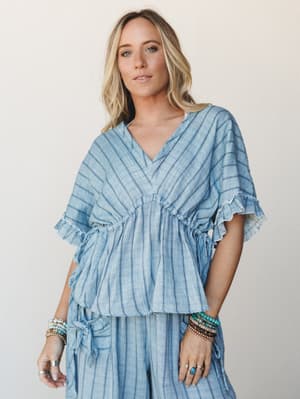 A breezy striped linen-look blouse with ruffle sleeve trim and a gathered waist, styled as a matching set with wide-leg pants and layered beaded bracelets — effortlessly relaxed with a coastal, sun-worn feel.