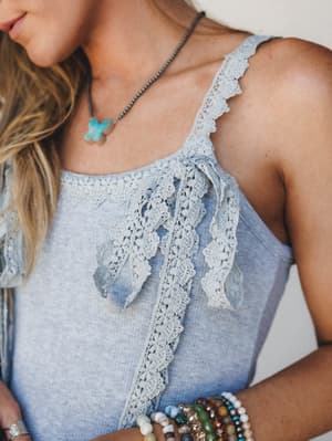 A close-up detail shot of a heather gray knit tank with delicate lace trim along the straps and neckline, featuring a lace bow tie at the center front — styled with layered bead bracelets and a turquoise butterfly pendant necklace for an easy, expres
