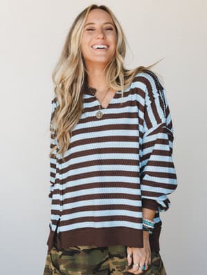 A laughing blonde model wears a relaxed waffle-knit striped pullover in mocha and sky blue, styled with camo pants and layered turquoise bracelets — the kind of effortless, feel-good outfit that just makes sense.