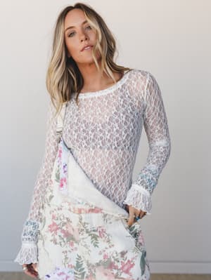 A sheer ivory lace long-sleeve top layered over a floral overall dress, styled with stacked rings and a relaxed hand-in-pocket pose that feels effortlessly romantic.
