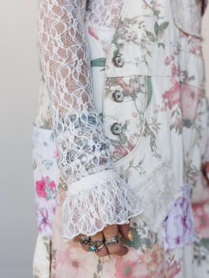 A close-up detail shot of an ivory floral top with sheer lace sleeves, silver concho buttons, and a lace ruffle cuff — the model's hand is stacked with silver and turquoise rings, adding a perfectly personal boho touch.