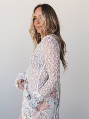 A model shot in soft studio light, wearing a sheer ivory lace long-sleeve top layered over a floral skirt — romantic and effortlessly undone, like she got dressed for something that mattered.
