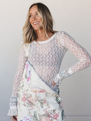 A smiling model layers a sheer ivory lace long-sleeve top over a floral overall dress, the combination feeling effortlessly romantic — like she got dressed by instinct and it all just worked.