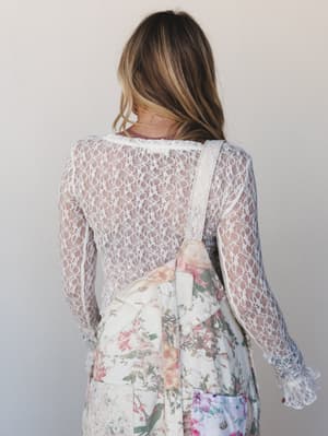 Shot from behind, a model layers a sheer ivory lace long-sleeve top over a floral slip dress, the delicate scalloped cuffs and open lattice lace pattern doing all the quiet talking.