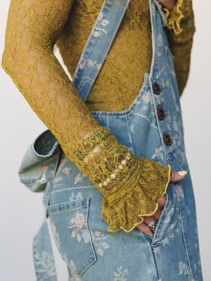 A close-up of a golden olive lace long-sleeve top layered under floral-printed denim overalls — the ruffled lace cuff and delicate floral denim together feel like something you'd find at a vintage market and never stop wearing.