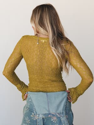Shot from behind, a model in a sheer olive-gold lace long sleeve top paired with a floral patchwork denim skirt — the kind of outfit that looks effortless but was absolutely thought about.