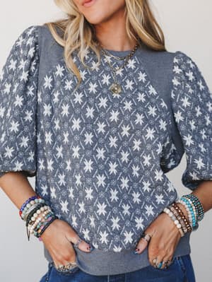 A slate blue patterned sweater top with puff sleeves styled with stacked beaded bracelets, layered gold necklaces, and denim — the kind of outfit that looks like you tried just enough.