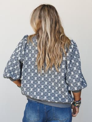A back-view shot of a quilted charcoal top with a repeating star and diamond print, puff sleeves, and ribbed hem — styled with denim and a stack of boho bracelets for an easy, expressive everyday look.
