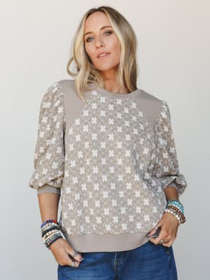 A relaxed pullover top in warm mushroom and white with a geometric star print, styled with stacked beaded bracelets and denim — the kind of top that looks intentional without trying too hard.
