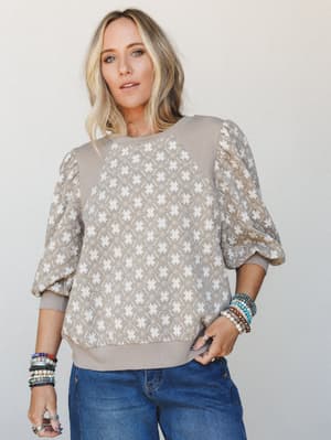 A relaxed puff-sleeve sweatshirt top in warm mushroom and cream with an allover geometric star print, styled with denim and stacked beaded bracelets — the kind of top that makes a regular Tuesday feel a little more intentional.