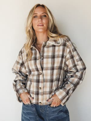 A plaid flannel shacket with a sweet lace collar detail — the kind of piece that makes a coffee run feel like a whole moment — worn over denim with hands tucked in pockets and a relaxed, faraway gaze.