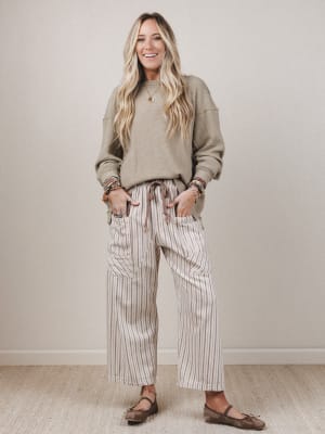 Woman wearing a beige sweatshirt and striped pants standing against a plain wall.