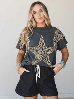 A washed charcoal graphic tee with a frayed leopard-print star appliqué and ruffle sleeves, paired with dark denim drawstring shorts and stacked turquoise and mixed-bead bracelets — the kind of effortless, personality-forward outfit that makes runnin