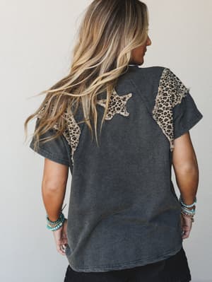A back-view model shot showing a washed ash tee with leopard print star and wing appliqués — the kind of top that feels effortless but has a little something going on, styled with stacked turquoise bracelets and black bottoms.