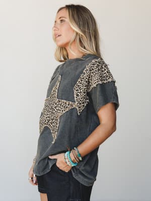 A washed ash charcoal graphic tee with a large leopard print star appliqué and frayed shoulder panel detail, styled with dark denim and a stack of turquoise and gold bracelets — the kind of top that looks effortless but is clearly doing a lot.
