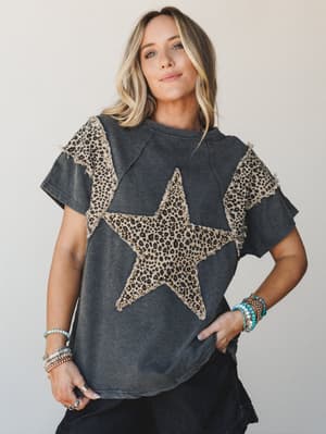 A washed ash graphic tee with a standout leopard-print star appliqué and raw-edge detailing — styled with stacked boho bracelets and dark jeans for that effortless, expressive everyday energy.
