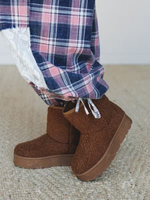 A close-up of cozy sherpa platform booties in warm chestnut brown, paired with plaid wide-leg pants and a peek of lace trim — the kind of detail shot that makes you want to immediately check the weather and justify buying both.