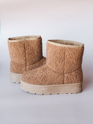 A pair of sherpa-textured platform booties in warm tan sit against a soft neutral backdrop — the kind of shoes that make you want to immediately put them on and never take them off.