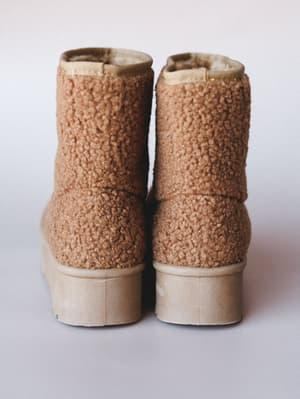 A pair of cozy sherpa ankle booties shot from behind, showing off their chunky platform sole and plush teddy texture in a warm camel tone — the kind of boots that make you want to walk slowly through fallen leaves.