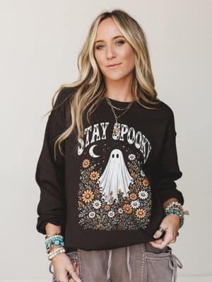 A dark chocolate graphic sweatshirt with a whimsical 'Stay Spooky' ghost-and-wildflowers print, styled with washed cargo pants and stacked boho bracelets — the kind of Halloween dressing that's more cottage witch than costume shop.