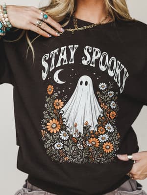 A close-cropped shot of a dark chocolate sweatshirt printed with a whimsical 'Stay Spooky' ghost-and-wildflower graphic, styled with layered silver and turquoise jewelry for an effortlessly boho Halloween vibe.