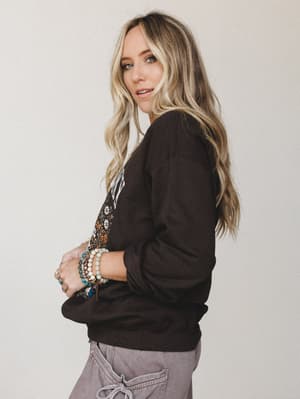 A model shot from the side with a over-the-shoulder glance, wearing a dark chocolate graphic sweatshirt with a floral and daisy motif, paired with washed grey cargo pants and a stack of boho beaded bracelets — the kind of outfit that feels just as go