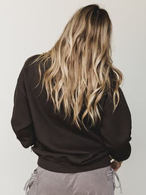 A back-facing shot of a model in a dark chocolate pullover sweatshirt paired with dusty lavender cargo pants — relaxed, unfussy, and the kind of outfit that makes a Saturday morning feel intentional.