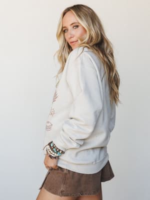 A sun-kissed blonde model glances back over her shoulder in a cream graphic sweatshirt layered over a brown mini skort, her wrist stacked with turquoise and pearl bracelets — the kind of effortless fall outfit that makes you want to light a candle an
