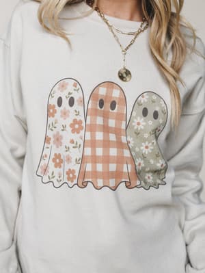 A cozy sand-colored pullover sweatshirt with a boho-cute ghost graphic — three little ghosts each dressed in their own floral or gingham print — styled with layered gold chain necklaces for that effortlessly warm, Halloween-with-a-soft-side vibe.