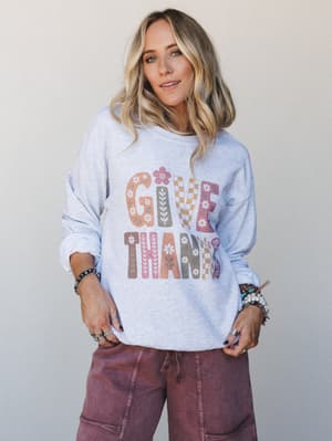 A relaxed ash grey graphic pullover with a boho-retro 'Give Thanks' print in mixed floral and checker patterns, styled with dusty mauve cargo pants and stacked bracelets — the kind of cozy-expressive combo you reach for on a crisp fall morning.