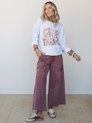 A relaxed full-body shot of a model in a heathered ash graphic sweatshirt paired with wide-leg mineral-washed plum pants and mixed sneakers — the kind of easy, expressive outfit that works from morning coffee to a laid-back Thanksgiving table.
