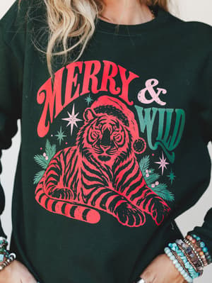 A forest green crewneck sweatshirt with a bold 'Merry & Wild' holiday graphic featuring a red illustrated tiger in a Santa hat, styled with stacks of beaded bracelets for a playful, boho-meets-Christmas vibe.