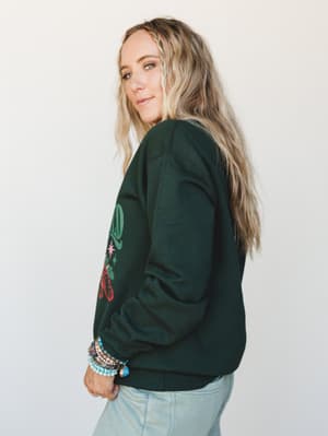 A side-profile shot of a woman in a forest green graphic crewneck paired with light wash jeans, her wrist stacked with beaded bracelets — the kind of cozy-cool combo that feels as easy as it looks intentional.