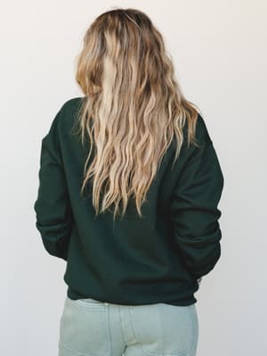A back-facing shot of a model in a deep forest green crewneck sweatshirt paired with light wash jeans — easy, lived-in, the kind of outfit that makes a coffee run feel intentional.