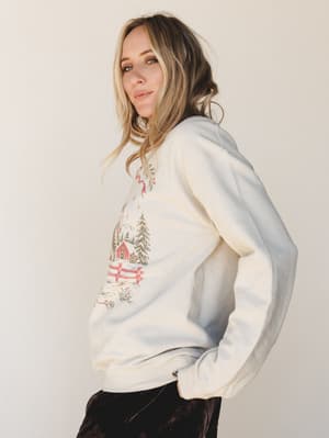 A cozy oversized crewneck sweatshirt in warm sand with a nostalgic winter countryside graphic — red barn, snow-dusted pines, and holiday details — worn with dark pants against a soft neutral studio backdrop, giving off that 'hot cocoa by the window'