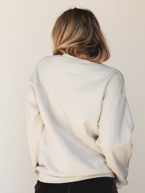 A relaxed cream crewneck sweatshirt shot from behind, styled with dark bottoms — the kind of top that makes a Sunday morning feel intentional.