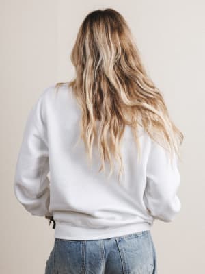 Person wearing a white sweatshirt and blue jeans with long blonde hair, standing against a plain background