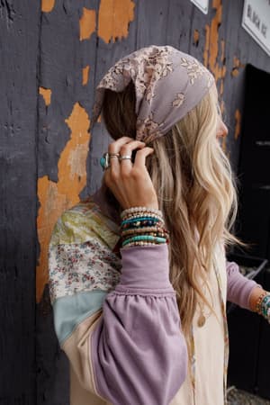A woman with long, wavy blonde hair stands in front of a worn wooden wall, wearing the Sarah Floral Lace Headscarf in Gray, a floral patchwork top, and beaded bracelets and rings. She looks away, gently touching her hair.