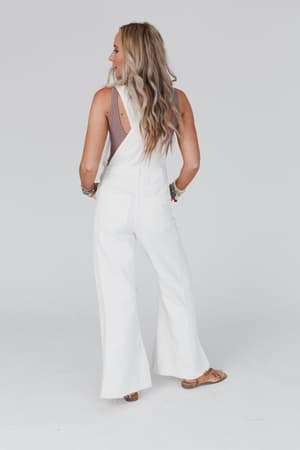 A woman with long, wavy blonde hair stands facing away in the Milly Distressed Hem Wide Leg Overall - Cream, paired with sandals and bracelets, set against a plain light background.